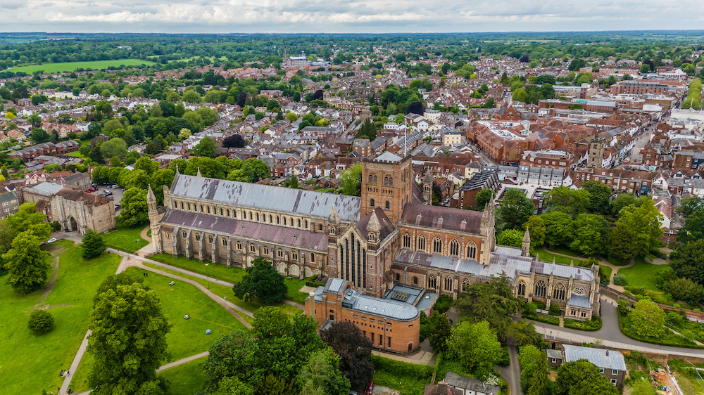 Aerial view of St Albans Cathedral rising above the city centre, with surrounding parkland, historic streets and the wider Hertfordshire landscape beyond.